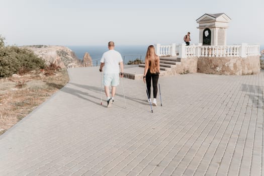 Happy Middle aged couple or friends practicing nordic walking in park near sea. Mature couple with trekking poles walking, practicing Nordic walking outdoors. Aging youthfully and sport concept.