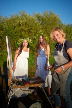 Adult female artist painting picture near water of river or lake in nature and girls in white sundress and flower wreath. Artist and models posing in holiday of Ivan Kupala in nature at sunset