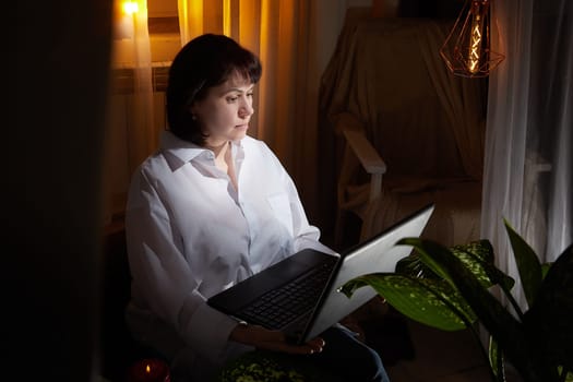 A girl in white shirt with a laptop in a dark room. A middle-aged woman works online remotely in the evening. The student is preparing for the exam at night. Blogger or freelancer in the chat