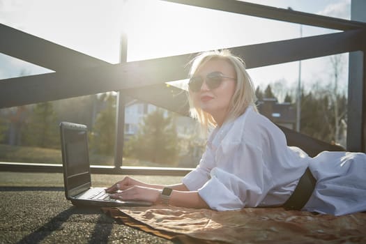 Fashionable beautiful blonde girl is working with laptop in gazebo on sunny autumn, spring day. Businesswoman, entrepreneur, student, manager works outdoors against the backdrop of natural landscape