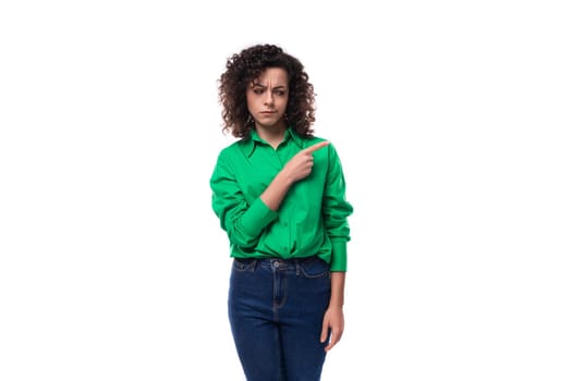 young caucasian brunette woman with curled hair dressed in a green shirt points her finger towards copy space. advertising concept.