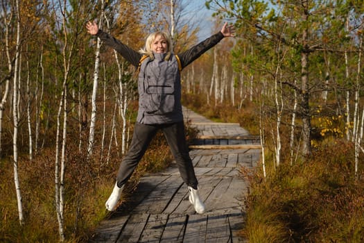 A happy woman with a backpack jumps on a wooden path in a swamp in Yelnya, Belarus.