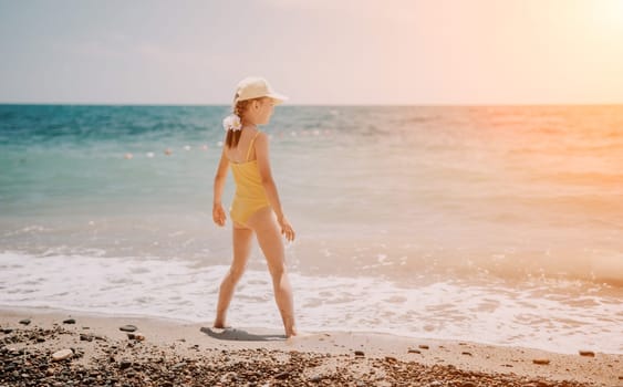 Cute little girl running along the seashore against a clear blue sea and rejoices in the rays of the summer sun. Beautiful girl in yellow swimsuit running and having fun on tropical beach