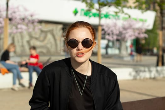 A girl in round glasses. Portrait of a young girl. The teenager looks through the glass of his sunglasses. A white girl with red hair.