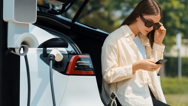 Young woman use smartphone to pay for electricity at public EV car charging station green city park. Modern environmental and sustainable urban lifestyle with EV vehicle. Expedient