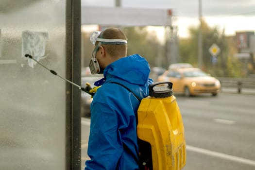 Guy washes bus stop. Worker with canister of water. Spraying detergent on glass. Laundering of urban infrastructure.