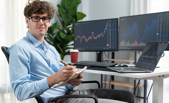 Profile of smiling young stock trader looking at camera, holding notepad and pen against on financial exchange rate screen at office. Concept of professional business digital investment. Gusher.