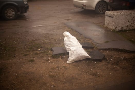 Bag with unknown contents on street. White bag stands on ground. Forgotten thing in parking lot. Smuggling of prohibited substances.