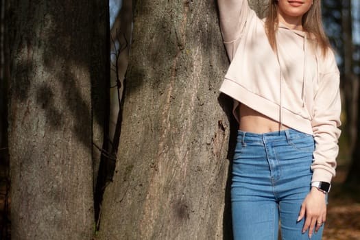 Girl stands by tree. Beautiful figure. Blue jeans. Young girl poses in park.