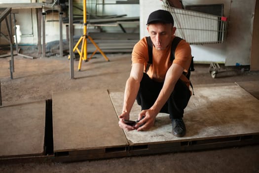 Guy is sitting on floor in garage. Guy looks at phone. Man is waiting for work. Man is squatting.