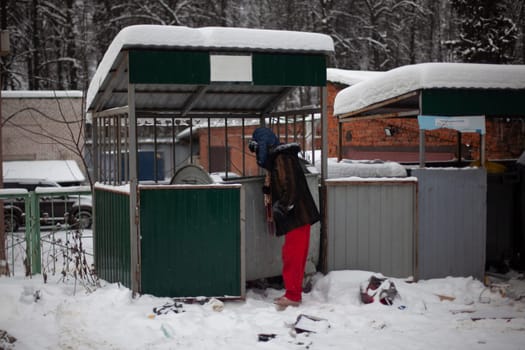 Man is looking for food in garbage. Man looks into dumpster in winter. Poor man on street. Garbage cans in Russia.