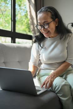 Happy middle age woman sitting on sofa chatting with friends in internet social networks.