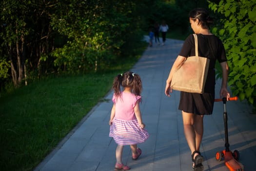 Mother with child walks through park. Walking with child in summer. Asian family on street. Mother carries her daughter's scooter.