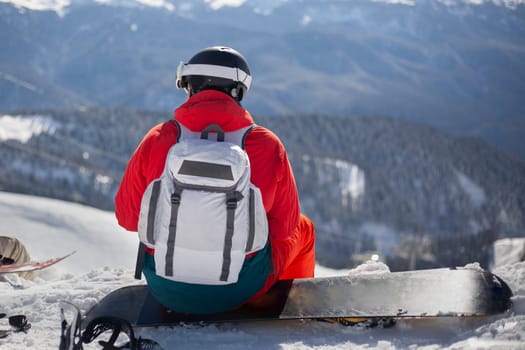 A snowboarder sits on his board on a snowy slope. A guy in a helmet to protect his head. The athlete is preparing for the descent from the mountain.