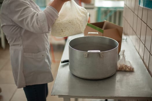 Cook prepares meals. Large pot on table. Cottage cheese in bag. Work as cook in dining room. White coat.