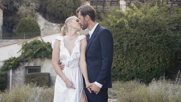 Newlyweds kiss and pose for camera. Action. Beautiful photo shoot of kissing couple of newlyweds. Wedding photo shoot of kissing newlyweds in garden.