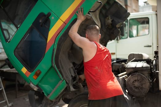 Driver repairs truck. Man lifts cab of truck. Big car. Guy in red T-shirt.