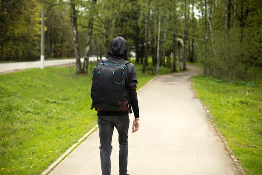 Guy walks through park in summer. Man in black clothes with backpack on his back. Student goes to school on street. Head in hood.