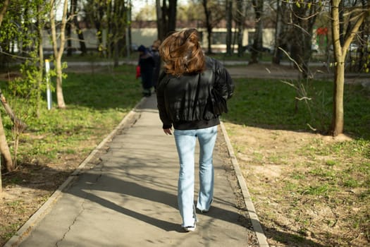 Girl walks down road. Woman in black jacket and blue jeans. Woman in park.