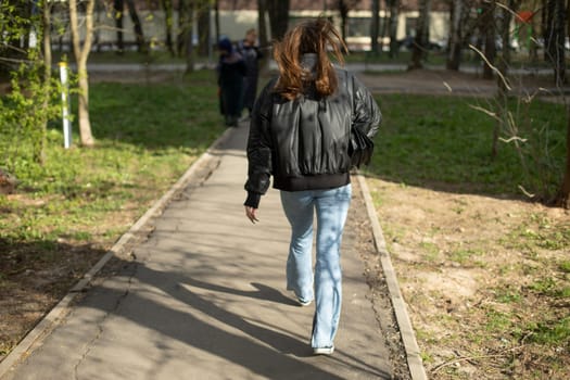 Girl walks down road. Woman in black jacket and blue jeans. Woman in park.