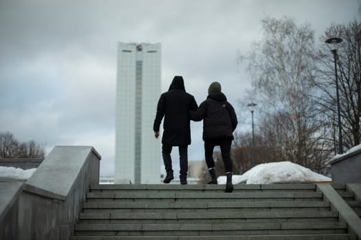 Guy and girl walk through city. Man leads girl under his arm. People on steps.