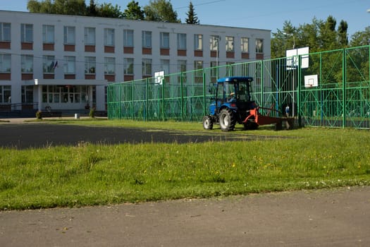 Tractor mows grass outside school. Cleaning of territory. Cutting grass in summer.