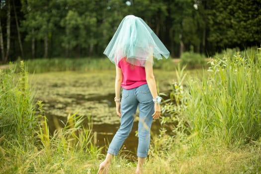 Girl in park. Girl in summer on shore of pond. Blue cape on head. Transparent veil on hair. Jeans and t-shirt.