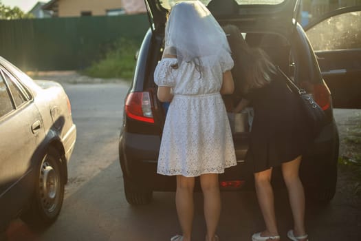 Bride is in parking lot. Bridesmaids at car dealerships. Trunk of car. Girls are getting ready for trip. Women in dresses.