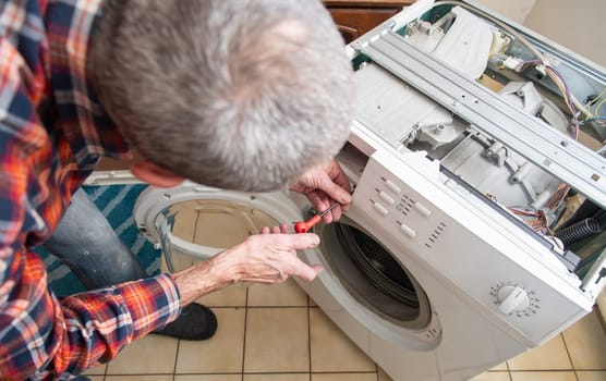 middle-aged master in a checkered suit unscrews the lid of a washing machine with a screwdriver for repair, repairs washing machines and household appliances at home, high quality photo