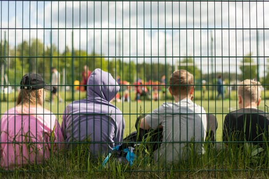 Children in stadium. Teenagers on sports field. Schoolchildren sit on grass behind net. Fence around football field. Spectators at sports competition.