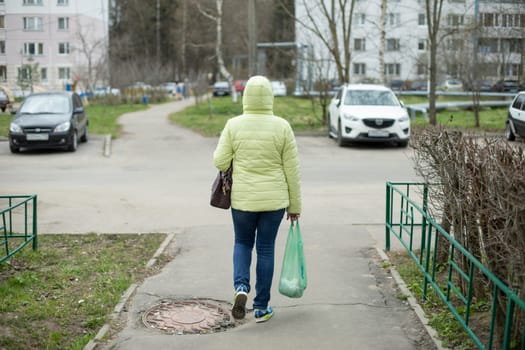 Woman walks with bag in her hand down street. Girl in green jacket. Person walks out of store with groceries in bag.