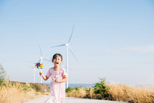 Little girl's happiness seen near windmills holding pinwheels while running. Playful wind energy education visuals showcasing clean electricity generation under a serene sky.