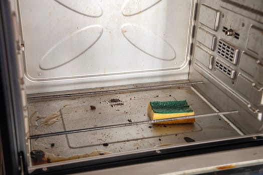 A yellow sponge with a cleaning solution lies in an open dirty oven with burnt grease stains on the internal surfaces, a freestanding oven on a table in the kitchen, high quality photo