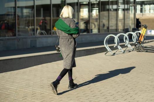 Girl in coat and with green scarf walks down street. Young lady walks around city. Girl with white hair.