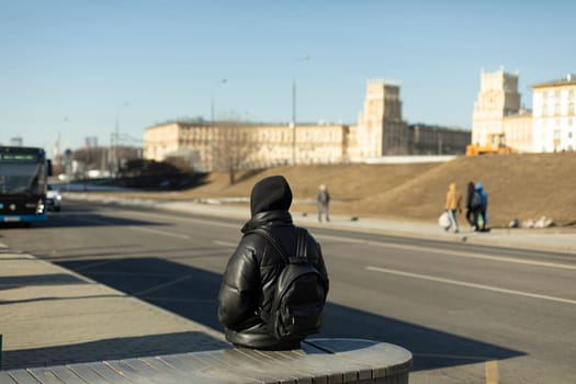 Guy is waiting for bus. Man in black clothes. Man is waiting for transport in city. Sitting on bench.