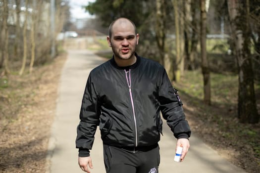 Guy in black jacket walks through park. Man on road in woods. Man hurries through park.