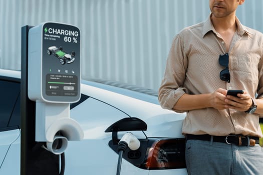 Young man use smartphone to pay for electricity at public EV car charging station at city commercial mall parking lot. Modern environmental and sustainable urban lifestyle with EV vehicle. Expedient