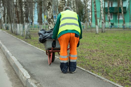 Worker carries cart of garbage. Work clothes of bright color. Cart with bags. Gardener cleans area.