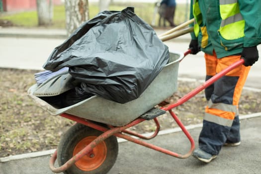 Worker carries cart of garbage. Work clothes of bright color. Cart with bags. Gardener cleans area.