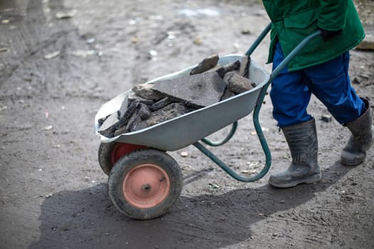 Builder removes broken stone. Man carries cobblestones in cart. Worker in green jacket makes embankment on road. Elimination of pits.