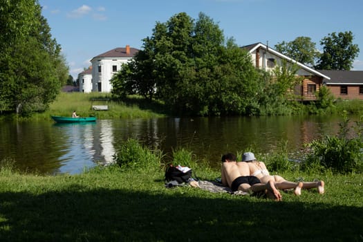 Beach on lake. Guy and girl lie on shore. Boat on water. Countryside outside city. Rest in summer.