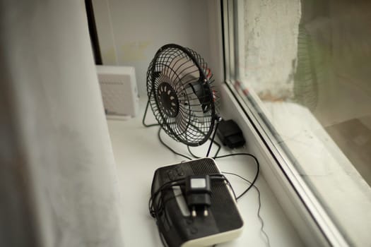 Fan is on window. Items lie on windowsill. Electrical appliances at home. Household appliances in apartment.