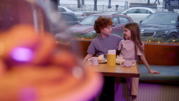 Cute little boy with curly red hair and a girl sitting together at the table cafe. Stock footage. Cookies and children enjoying cakes and drinks