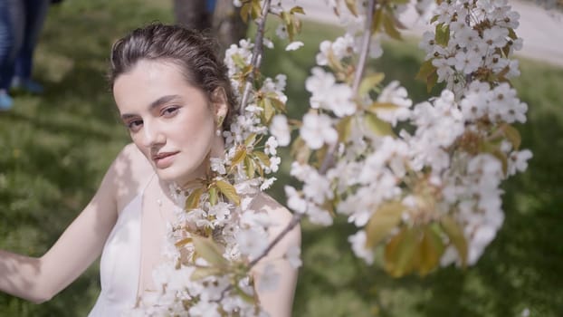 Woman posing with flowers of apple tree. Action. Thoughtful young bride in gorgeous wedding dress holding holding a branch of apple tree in a summer garden
