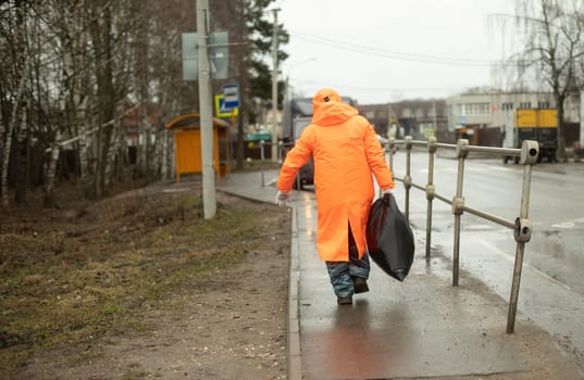 Worker removes garbage from road. Man in orange rain. Black bag in his hand. Road worker.