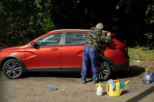 Man washes red car. Pensioner washes car. Work with transport. Detergents near car.