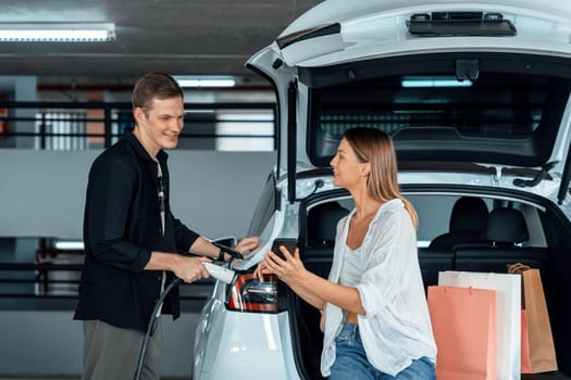 Young couple travel with EV electric car to shopping center parking lot charging in downtown city showing urban sustainability lifestyle by green clean rechargeable energy of electric vehicle innards