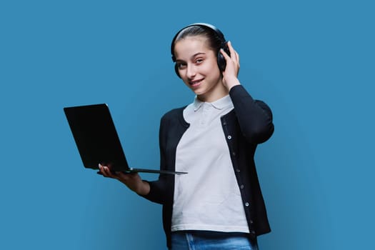 Teen girl student wearing headphones using laptop, looking at camera on blue studio background. Audio video internet technologies in education learning, online lessons, adolescence high school concept