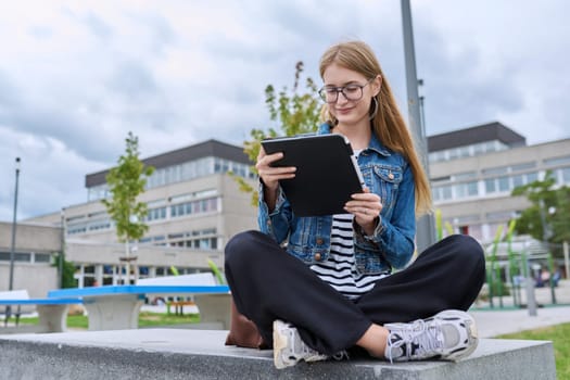 Girl student teenager outdoor near school building. Smiling teenage female with backpack, looking at screen of digital tablet. Adolescence, education, learning concept