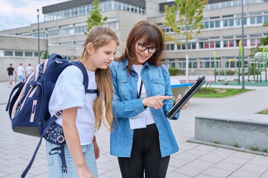 Talking female teacher and schoolgirl child outdoor, school building background. Meeting communication student girl with backpack and mentor counselor. Education, pre-teenage, learning, back to school